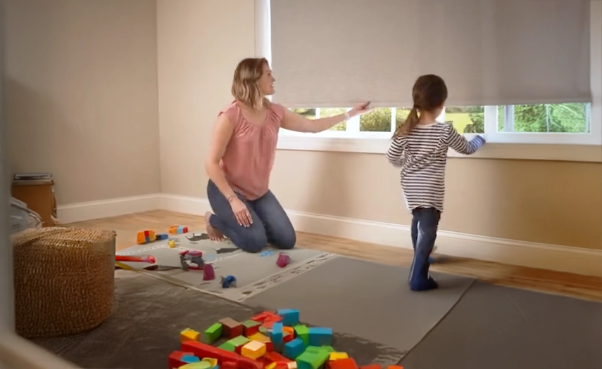 Mother adjusting window shade while her child plays.